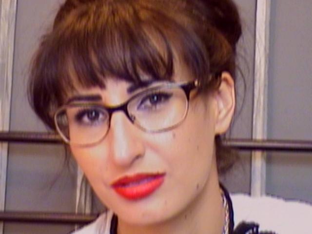 LysAddiction has dark brown hair in a bun, wears glasses, and has bright red lipstick on her face with a small beauty mark below her right eye.
