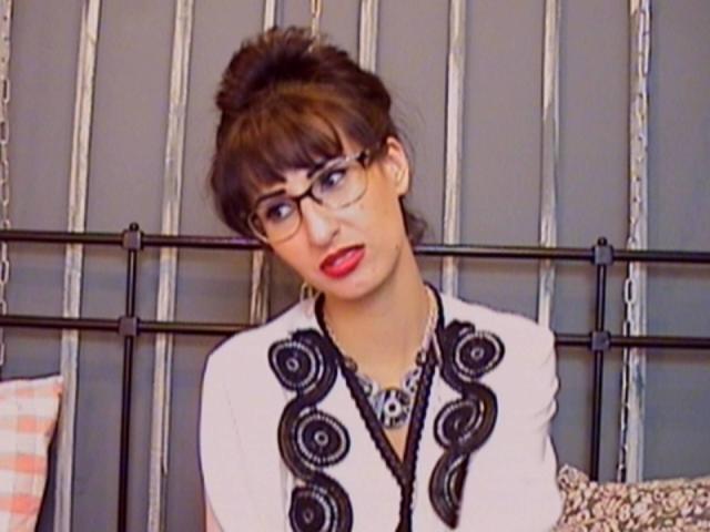 LysAddiction with glasses, red lipstick, and brown hair in a bun, wearing a white top with black swirls, looking slightly to the side.