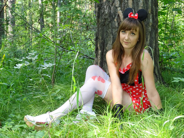 MilagroBest sentada na grama, vestindo um traje da Minnie Mouse com um vestido vermelho com bolinhas, meias brancas com laços vermelhos, luvas pretas e orelhas pretas com laço vermelho. Seios visíveis..