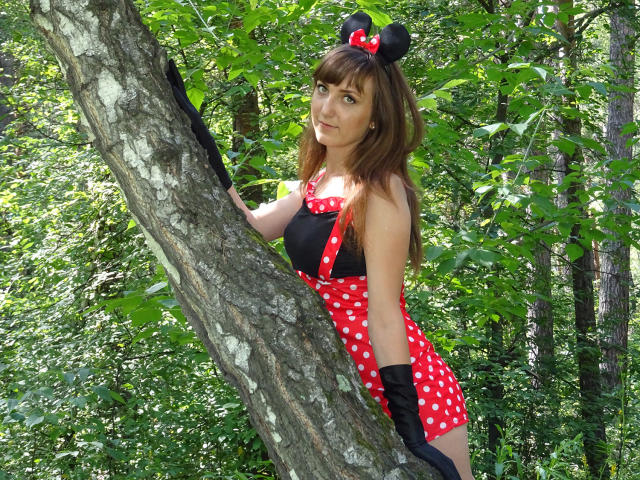 MilagroBest, gekleidet in einem roten, gepunkteten Minnie Maus Outfit mit schwarzen Handschuhen, lehnt an einem Baum in einem Wald und trägt Mäuseohren mit einer roten Schleife..