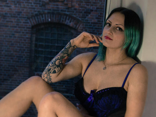 Erin, con cabello negro con puntas verdes, sostén de encaje negro, brazo tatuado, lápiz labial rojo, sentada, lazo morado, collar, mirando a la cámara, fondo de ventana, sexy, segura de sí misma, linda..