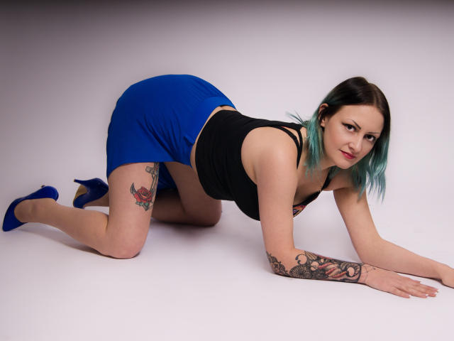 ErinHotBody è in posizione a quattro zampe, indossa una maglietta nera e una gonna blu, con tacchi azzurri, mostrando tatuaggi sulla coscia e sul braccio, e guarda verso la fotocamera..
