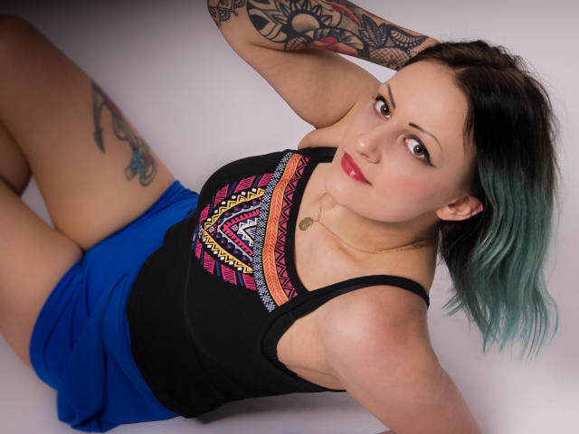 ErinHotBody mit türkisfarben gefärbtem Haar, schwarzem Tanktop und blauen Shorts, mit sichtbaren Tattoos, liegend, lächelnd, mit rotem Lippenstift und einer kleinen goldenen Halskette. Brüste sichtbar, Arm erhoben..