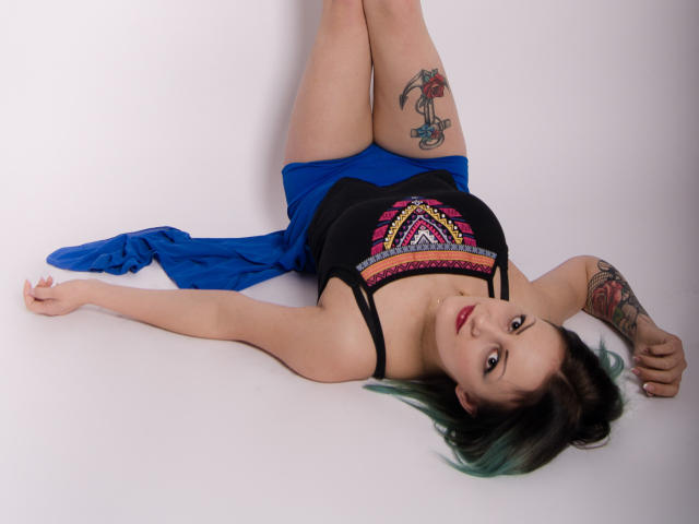 ErinHotBody ligt op haar rug met haar benen omhoog, een zwarte tanktop, een blauwe rok, tatoeages op haar dij en arm, groen geverfd haar, rode lipstick, en kijkt naar de camera..