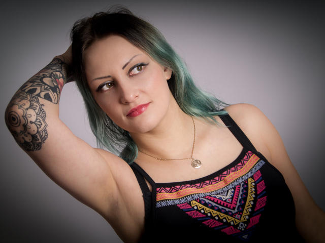 Erin, con cabello azul turquesa, camiseta negra, patrones geométricos coloridos, tatuaje en el brazo, mano en el cabello, collar dorado, lápiz labial rosa, sonrisa segura, iluminación suave, fondo gris..