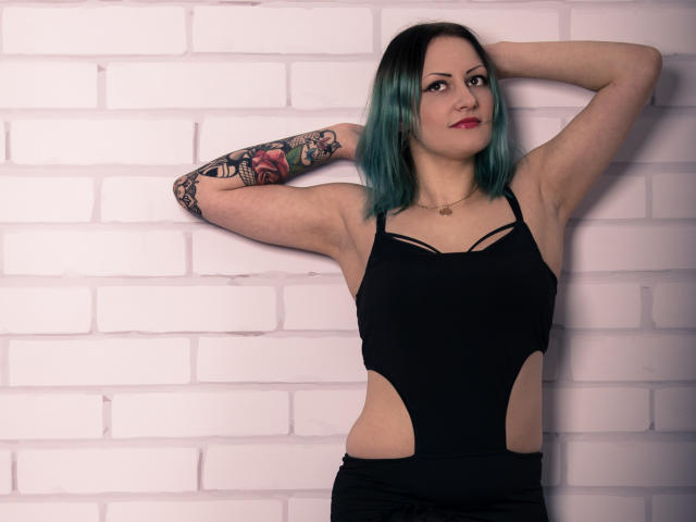 Erin, con cabello teñido de color turquesa, vestido negro con aberturas, brazo tatuado, brazos levantados, de pie frente a una pared de ladrillos blancos, con una ligera sonrisa, lápiz labial rojo, pose segura..