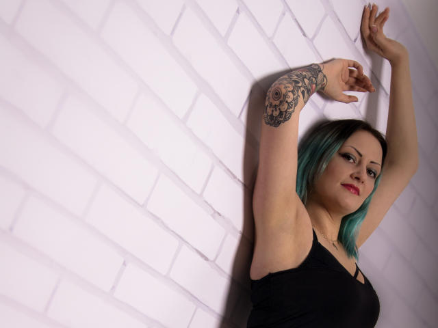 Erin, avec des cheveux teints en bleu turquoise, un débardeur noir, les bras levés, montrant un tatouage sur le bras droit, du rouge à lèvres et un léger sourire, devant un mur de briques blanches..