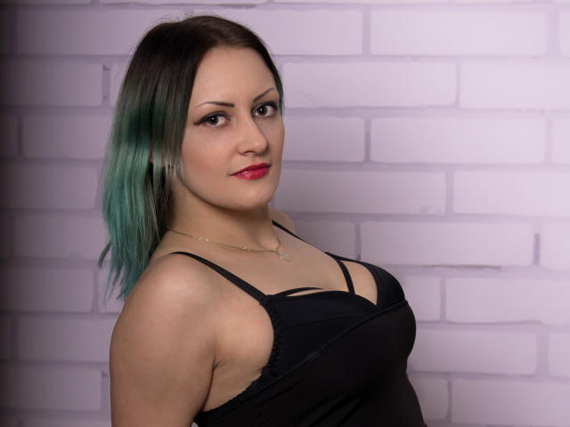 ErinHotBody mit türkisfarben gefärbten Haaren, rotem Lippenstift, schwarzem Kleid und kleinen Brüsten, blickt in die Kamera, lächelt, trägt eine goldene Halskette und steht vor einer weißen Ziegelwand..