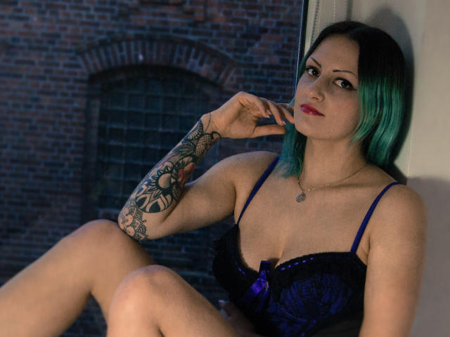 Erin, con cabello negro con puntas verdes, un sujetador de encaje púrpura, un brazo tatuado, lápiz labial rojo y un collar, sentada junto a una ventana, mirando a la cámara, con un dedo en la mejilla..