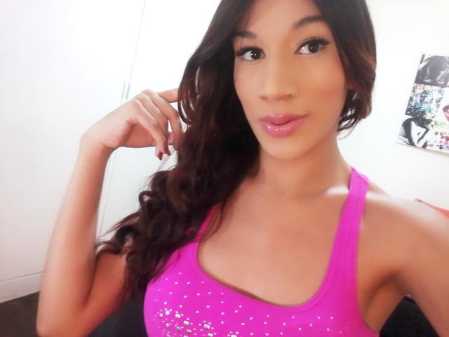 Littlebrunettehot with shiny pink lips, wearing a sparkly pink tank top, showing her boobs, long wavy brown hair, and a cute smile, with her hand near her face.