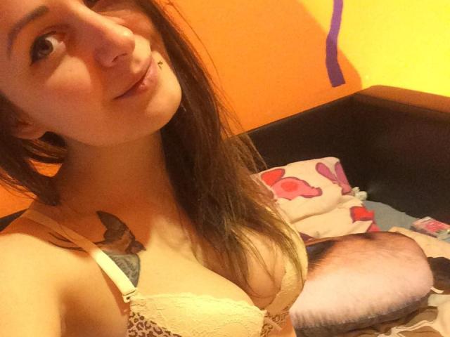 LounaDiva with long brown hair, yellow lace bra, small tattoo on left chest, and nose ring, sitting on bed with fluffy pillow and colorful blanket. Boobs visible.