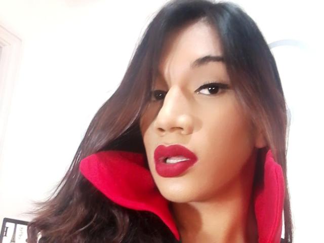 Una ragazza moro, con rossetto rosso, capelli castano scuro ondulati e un colletto rosso su una camicia nera, che guarda seducentemente la fotocamera..