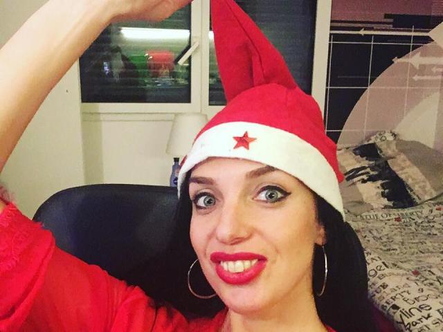 KatieFrenchie with red Santa hat, white trim, red star, and red lipstick, smiling, big green eyes, hoisting hat, red shirt, black hair, hoop earrings, bed in background.