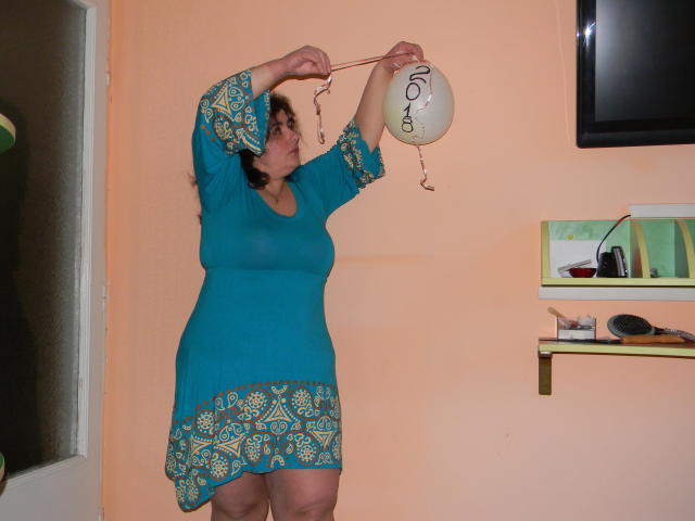 Keyx, une femme aux formes généreuses avec une poitrine de taille moyenne, portant une robe turquoise ornée de motifs jaunes, accroche une lanterne avec une écriture calligraphique, se tenant dans une pièce de couleur pêche..