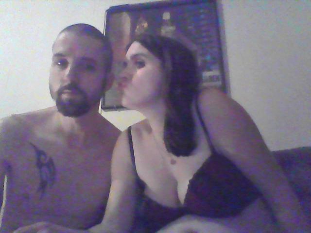 Un homme noir, torse nu, avec une barbe et un tatouage, est assis à côté d'une femme blanche aux cheveux foncés, qui l'embrasse, portant un soutien-gorge noir et montrant ses seins..
