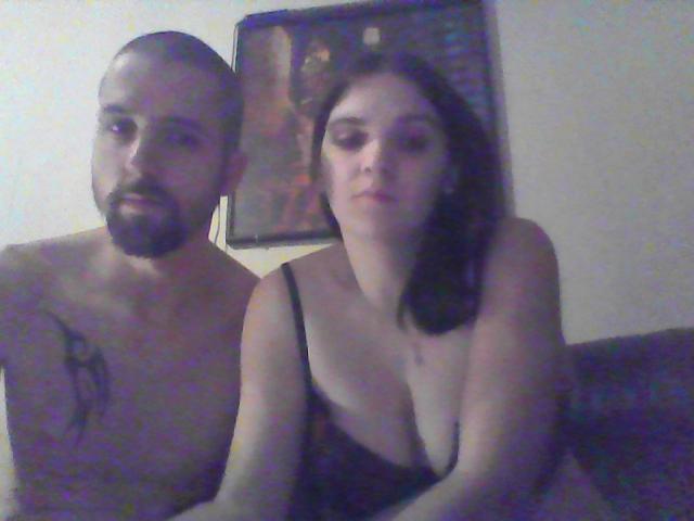 Shirtless SexiCouple with a tattoo on the left has a serious face, while the right SexiCouple with big tits and a black strap dress looks bored.