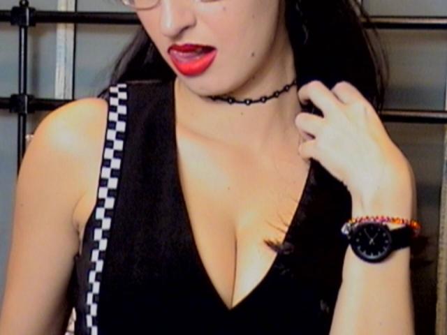LysAddiction with red lipstick, black choker, glasses, black sleeveless top showing big tits, checkered strap, black wristwatch, hand touching neck, dark hair.