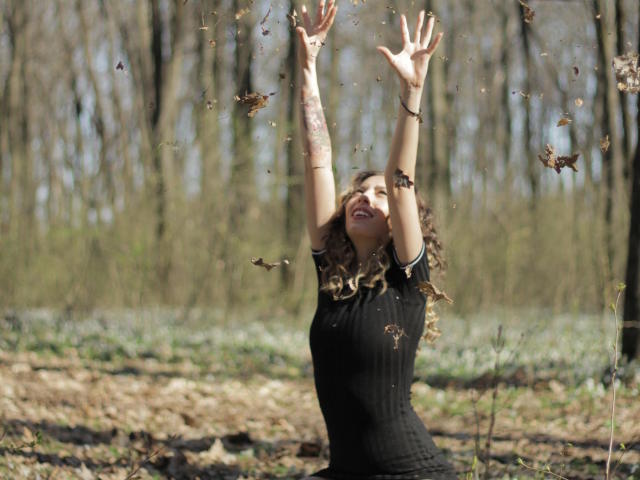 PoxyVibe with curly blonde hair, arms raised, black dress, surrounded by flying brown leaves, smiling, forest background, spring, sunlight, tattoos on forearms.
