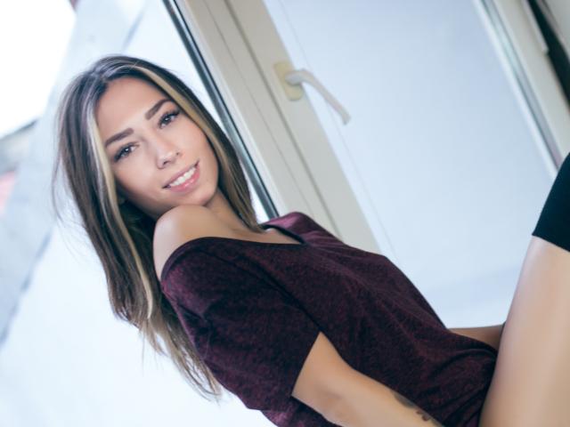 PoxyVibe has long brown hair, a cute smile, and wears an off-shoulder maroon top, sitting by a window, showing her bare ass in black panties.