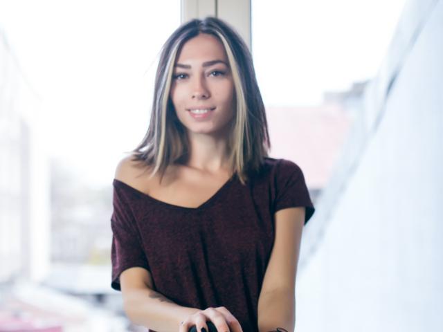 PoxyVibe has a cute smile, medium tits, and is wearing a maroon off-shoulder top, standing near a bright window with blonde hair and a tattoo on her right arm.
