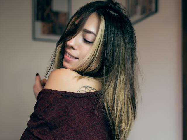 PoxyVibe with blonde and brown hair, off-shoulder maroon top, tattoo on right shoulder, smiling, eyes closed, hand near collarbone, soft lighting, relaxed, cute, attractive.