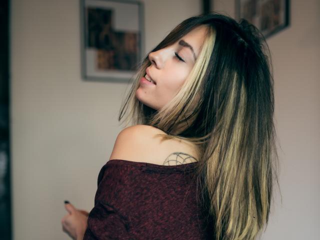 PoxyVibe with blonde and brown hair, eyes closed, smiling, wearing a red off-shoulder top, showing a tattoo on her left shoulder. She's pointing with her right hand.