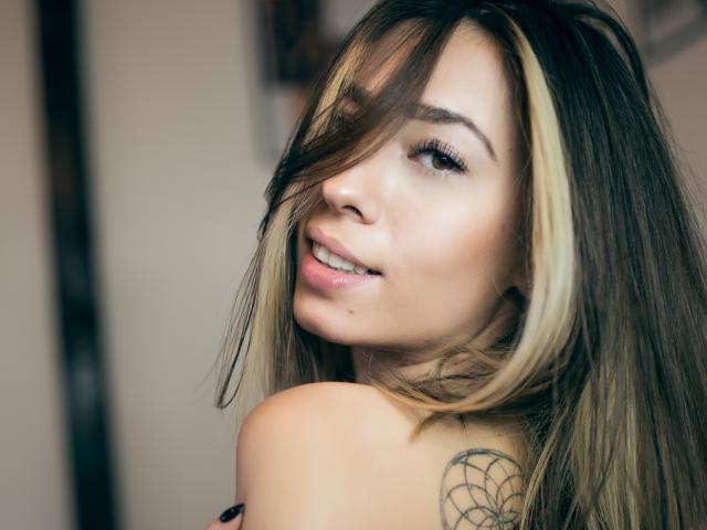 PoxyVibe has blonde and brown hair, a dreamcatcher tattoo, and is smiling with her ass partially visible, looking over her shoulder. She has light skin and eyeliner.