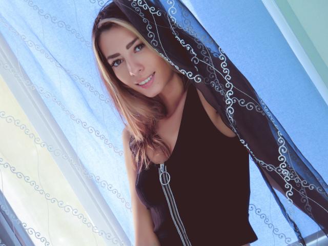 PoxyVibe with blonde hair, blue eyes, wearing a black tank top, standing in front of a sheer black curtain with white patterns, smiling softly at the camera.