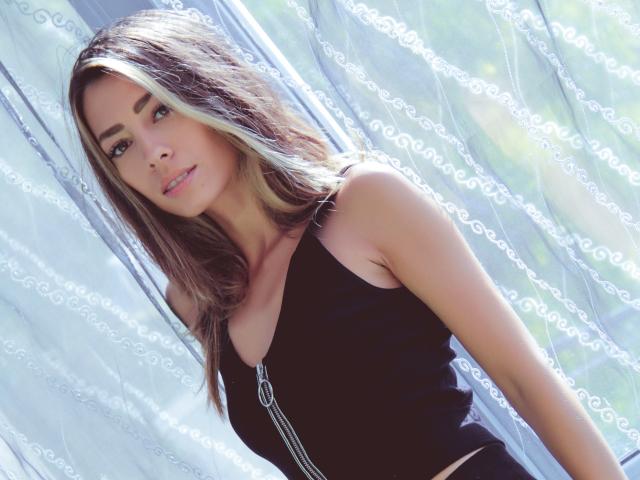 PoxyVibe with long brown hair and blonde highlights, wearing a black top with a zipper, has small tits and a cute face, standing in front of sheer curtains.