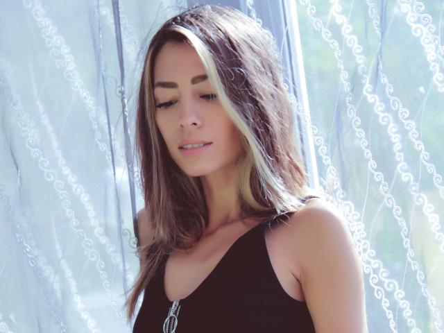 PoxyVibe has brown hair with blonde highlights, wearing a black top, looking down with a slight smile, standing in front of sheer, white curtains.