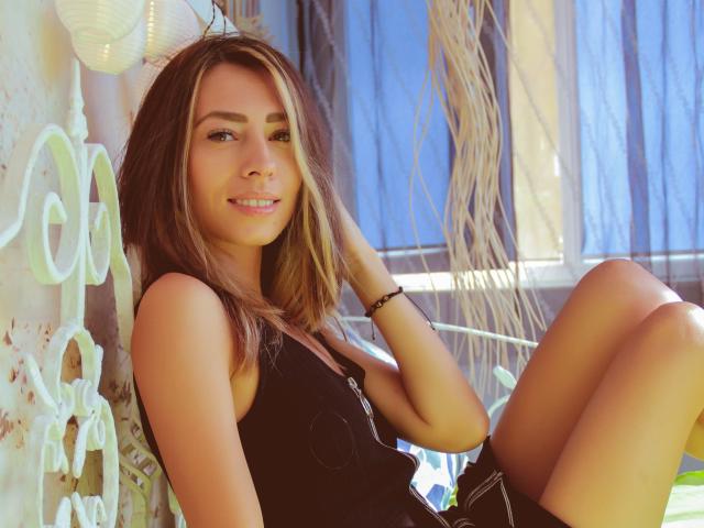 PoxyVibe with brown hair, blue eyes, and tan skin, wearing a black tank top and shorts, sitting with a hand in her hair, smiling, in a sunlit outdoor setting.