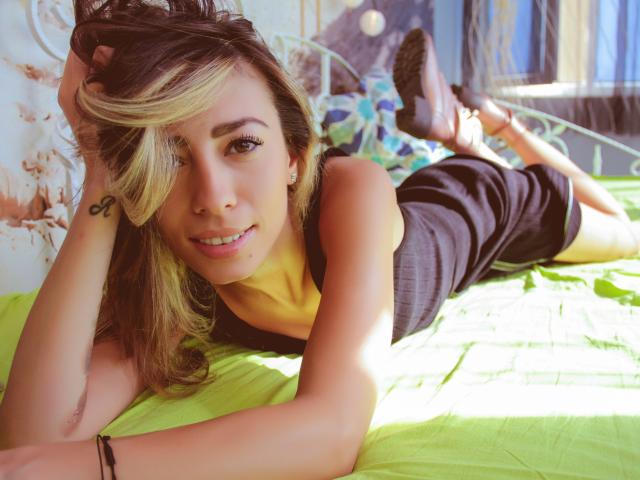 PoxyVibe with blonde and brown hair, lying on a green bed, wearing a black top, smiling, hands in hair, tattoo on wrist, green blanket, black boots, sunlight.