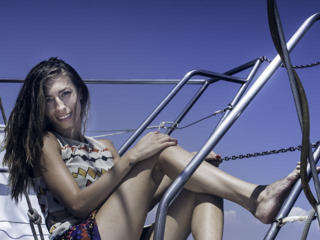 PoxyVibe has wet brown hair, wearing a zigzag-patterned top, sitting on metal ship stairs with legs up, barefoot, smiling, against a clear blue sky. Boobs are small.
