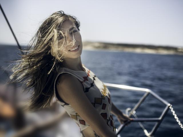 PoxyVibe with flowing brown hair, smiling, wearing a sleeveless, patterned top, holding a boat's metal rail, standing on a boat in a sunny, blue-water setting.