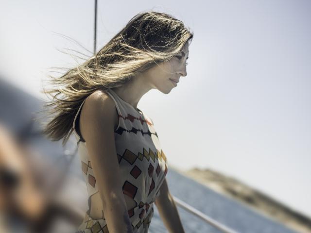PoxyVibe with blonde hair, wearing a geometric-patterned sleeveless top, stands in profile, sunlight and wind in her hair, smiling slightly. Boobs visible under top. Soft, natural light.