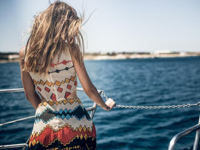 PoxyVibe's ass is facing the camera, wearing a colorful, patterned dress, standing on a boat with blue water and a metal railing, wind blowing through her blonde, wavy hair.