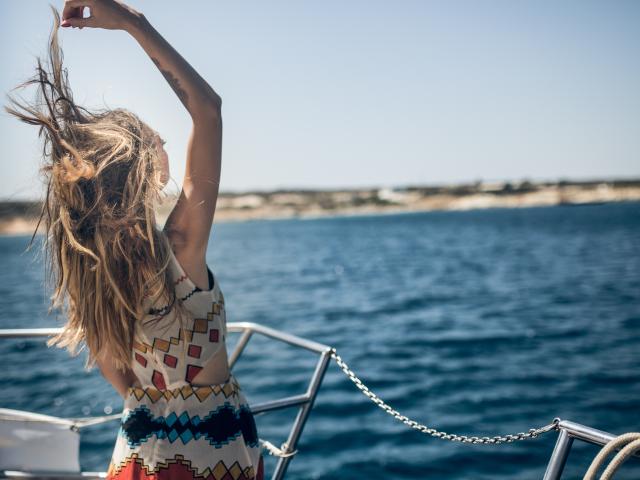 PoxyVibe with long blonde hair flipping it up, wearing a colorful geometric dress, standing on a boat with blue water and metal railings in the sunny background.