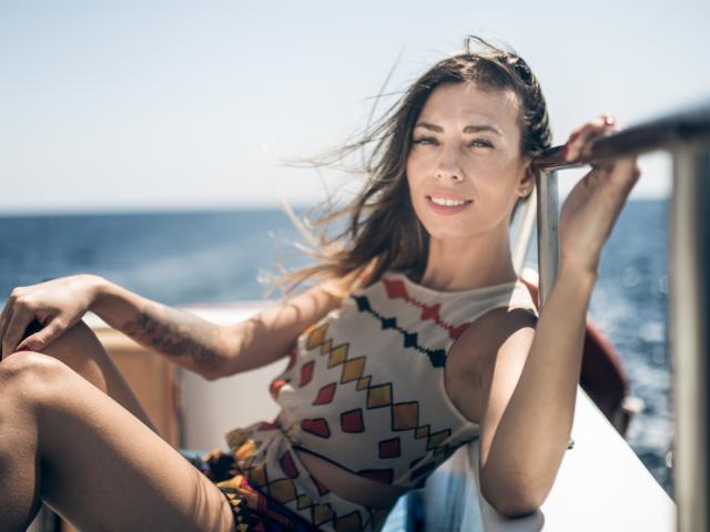 PoxyVibe with sunlit brown hair, smiling, wearing a white, geometric-patterned top, holding her hair, showing tattoo on left forearm, sitting on a boat, with blue ocean background.