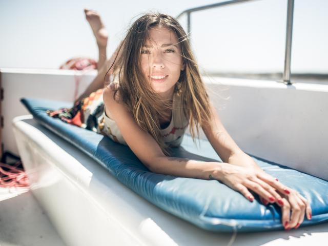 PoxyVibe lies on a blue inflatable mattress, sunlit, with long brown hair, a smile, and red nails, wearing a floral tank top, on a boat.
