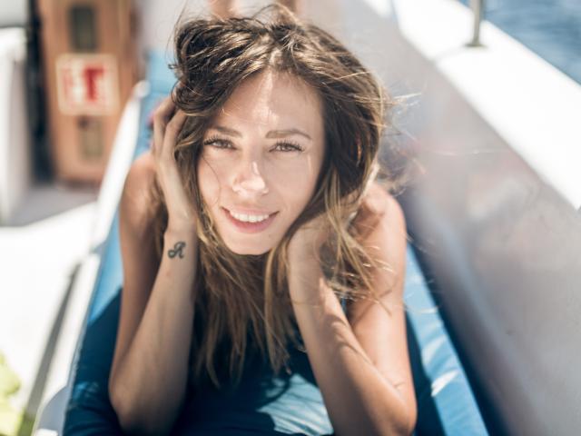 PoxyVibe has long brown hair, blue eyes, and a tattoo on her right wrist, smiling with hands in her hair, lying in a blue boat. Sunlight highlights her face.