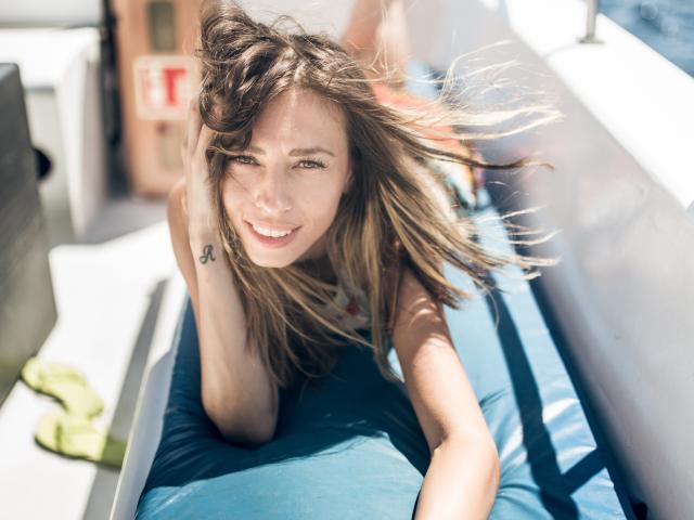 PoxyVibe, with dirty blonde hair flying, smiles brightly while lying on a blue cushion, showing a small tattoo on her right arm, and has a slim, attractive face.