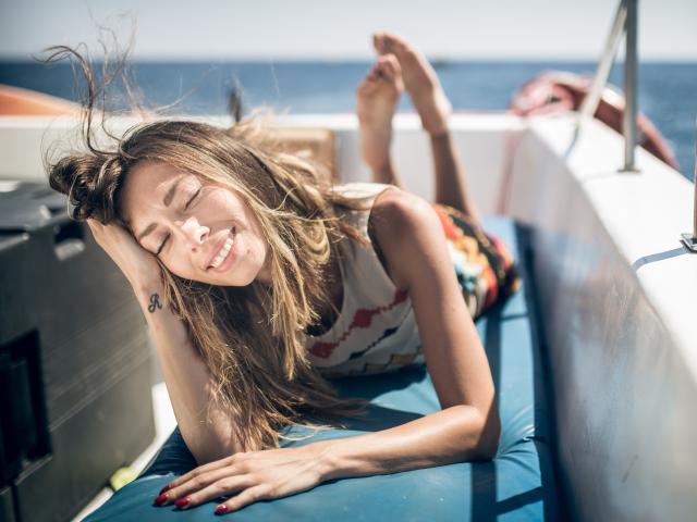 PoxyVibe's tits are small, she's lying on a boat's blue seat, smiling with eyes closed, wearing a colorful patterned top, and her long brown hair is blowing in the wind.