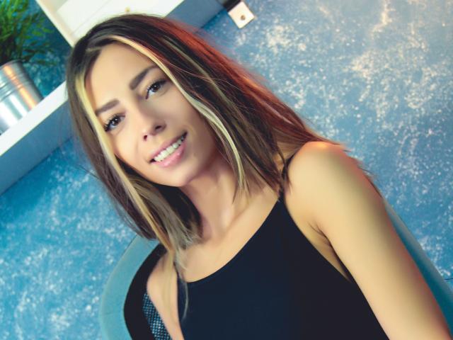 PoxyVibe with blonde highlights, smiling, wearing a black tank top, showing her tits in a blue-patterned background, sitting with a relaxed, sexy vibe.