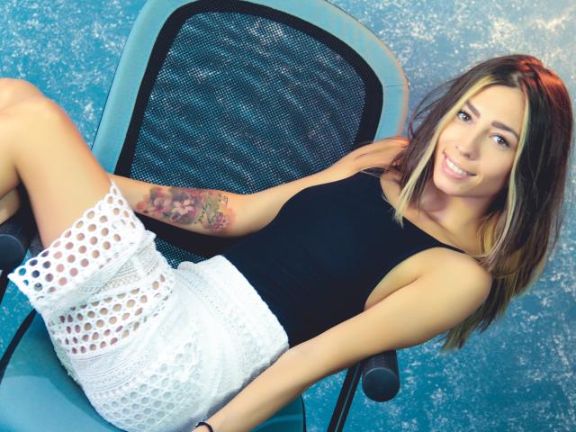 PoxyVibe, with blonde highlights, wearing a black tank top and white polka-dot skirt, has a colorful tattoo on her right arm, sitting on a chair, smiling.