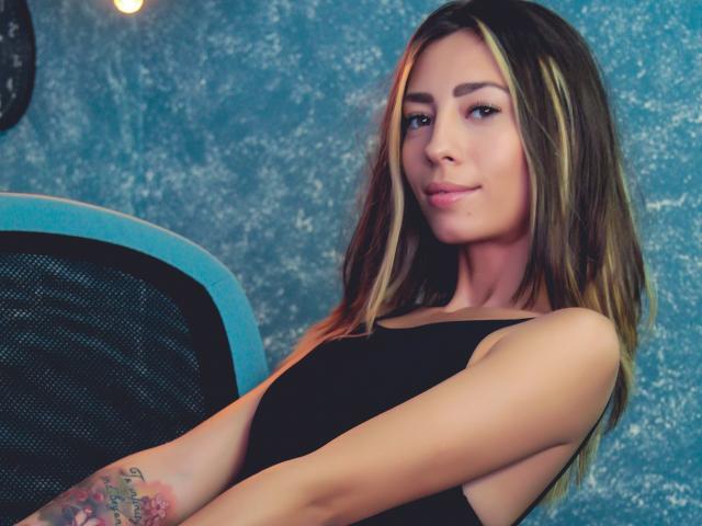 PoxyVibe with light brown hair, black tank top, tattoo on left forearm, sitting, smiling, blue textured wall, black mesh chair, warm light in background.