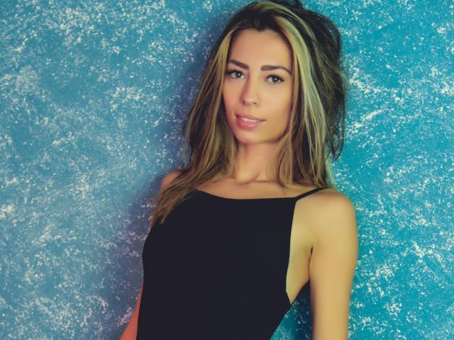 PoxyVibe with long blonde hair, black sleeveless top, and cute smile, standing against a blue textured wall, showing small tits and a slim, attractive body.