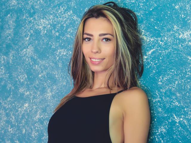 PoxyVibe with blonde and brown hair, smiling, wearing a black sleeveless top, showing her tits, against a textured blue wall. She's cute with brown eyes.