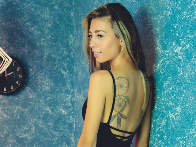 PoxyVibe with blonde hair, smiling, back to camera, black strap top, showing a large floral tattoo on her back, standing against a textured blue wall.