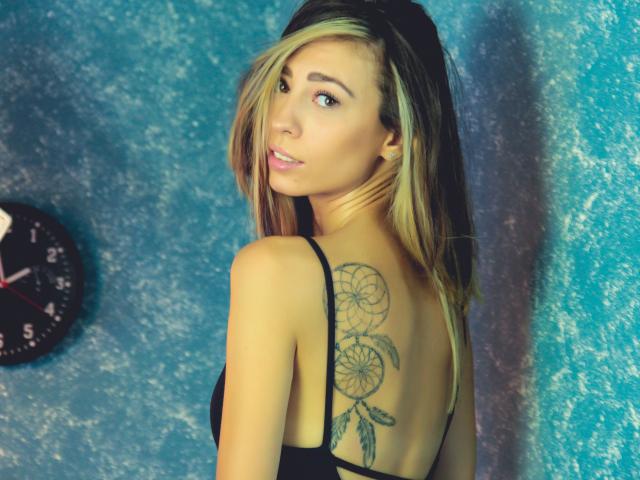 PoxyVibe with blonde hair, black strappy top, showing a dreamcatcher tattoo on her back, looking over her shoulder, blue textured wall, clock on the left.