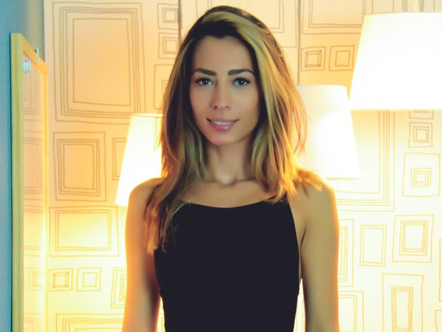 PoxyVibe with long blonde hair, wearing a black sleeveless dress, smiling at the camera, standing between two lit lamps, glowing softly. PoxyVibe's tits are subtle, her ass is not.